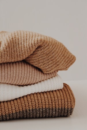 A neatly stacked pile of knitted sweaters in various shades, including cream, tan, and brown. The texture of the fabric is clearly visible, suggesting warmth and coziness.