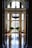 A grand interior space with a high ceiling and large arched windows allowing natural light to illuminate the polished wooden floor. An ornate chandelier hangs from the ceiling. Elegant tables and chairs flank the room, and lush green plants add a touch of nature near the windows.