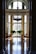 A grand interior space with a high ceiling and large arched windows allowing natural light to illuminate the polished wooden floor. An ornate chandelier hangs from the ceiling. Elegant tables and chairs flank the room, and lush green plants add a touch of nature near the windows.