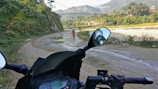 A motorcycle is parked on a muddy road with a scenic view in the background. A person wearing colorful attire is walking along the path, bordered by lush green hills and distant mountains. A river flows to the side, and a bridge can be seen in the distance under a clear sky.