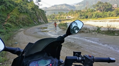 A motorcycle is parked on a muddy road with a scenic view in the background. A person wearing colorful attire is walking along the path, bordered by lush green hills and distant mountains. A river flows to the side, and a bridge can be seen in the distance under a clear sky.