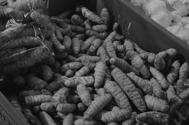 A pile of turmeric roots fills a container alongside some garlic bulbs on the side. The turmeric is arranged in a disordered manner, with some wrapped in plastic and others loose.