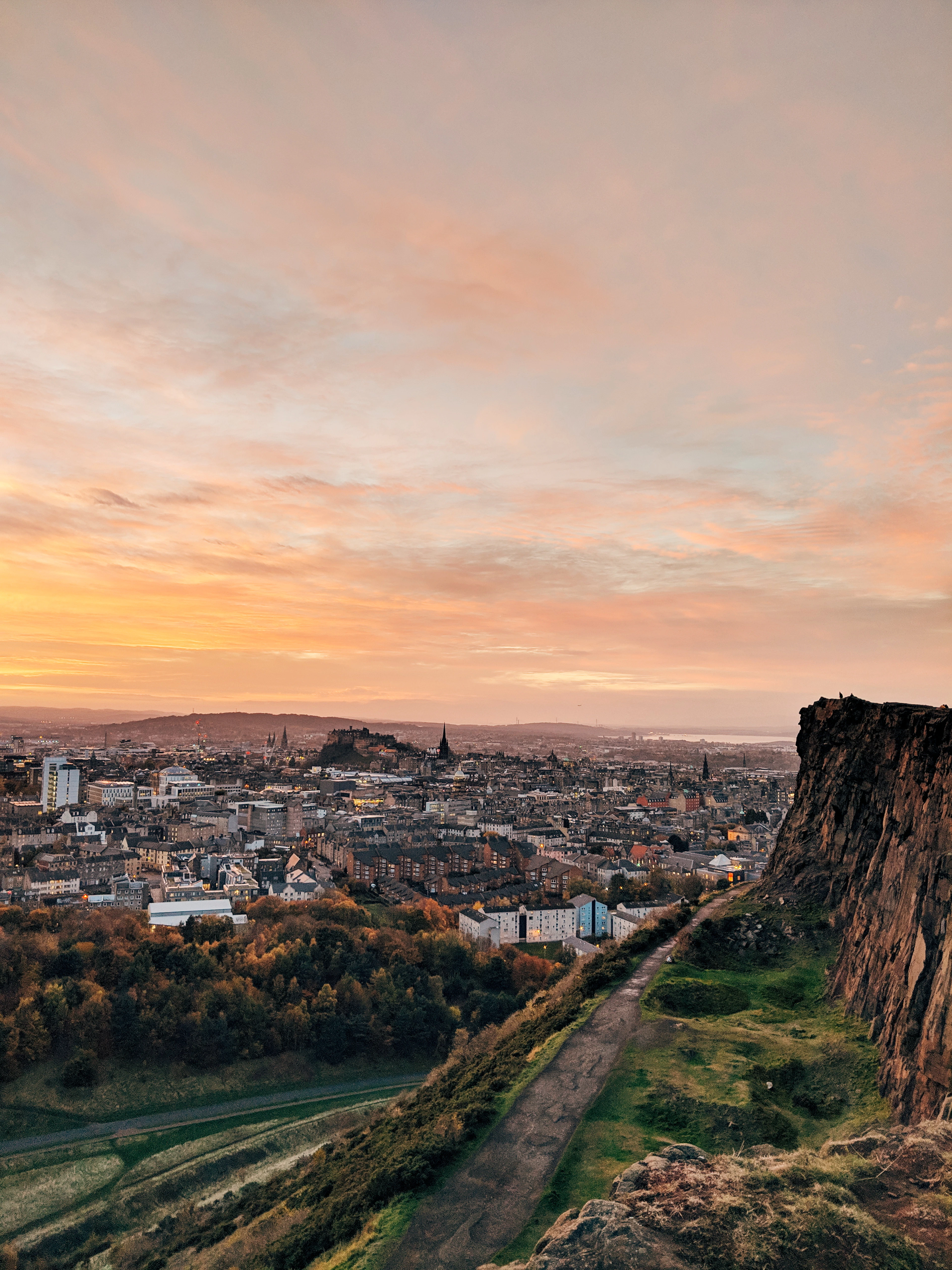The sheer dolerite cliff of Salisbury Crags in Edinburgh with the Radical Road path curving along its base, the whole city and its castle laid out below under a pink and gold sky.