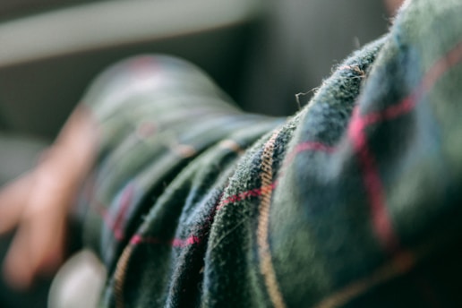 A close-up view of a plaid fabric with a soft focus, showing green as the primary color with accents of red and beige intersecting lines. The fabric appears to be part of a sleeve, with a blurred hand visible in the background.