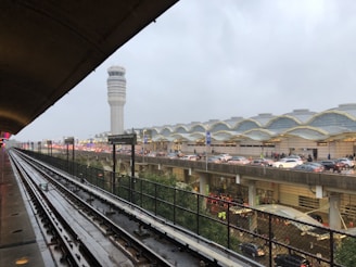 A train station platform overlooking a busy airport with a distinctive control tower. The scene is overcast with grey skies. The airport building has an arched roof, and there is a congestion of cars on the roads leading to the terminal.