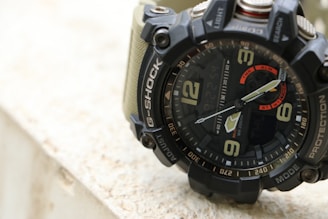 A close-up view of a rugged analog-digital wristwatch with a black bezel and bold numerals. The watch features multiple dials and buttons for various functions, including light and mode controls. The strap appears to be made of a sturdy material, possibly resin or fabric, and is olive green in color.