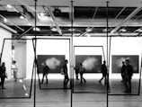 A group of people walk through an art gallery, where monochrome paintings are displayed on the walls. The perspective includes geometric frames hanging from the ceiling, adding a modern artistic effect. The setting is industrial with visible ceiling beams and lighting fixtures.