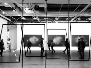 A group of people walk through an art gallery, where monochrome paintings are displayed on the walls. The perspective includes geometric frames hanging from the ceiling, adding a modern artistic effect. The setting is industrial with visible ceiling beams and lighting fixtures.