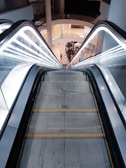 Close-up of an escalator handrail and steps in a busy shopping mall.