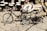 A collection of traditional drums and colorful painted musical instruments is displayed on a cobblestone surface. Two small decorative bicycles are positioned in the foreground, with intricate designs and patterns adorning their frames. The background is filled with various types of drums, including some with vibrant motifs and designs.