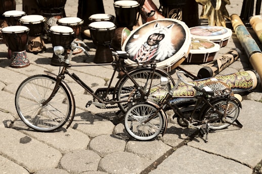 A collection of traditional drums and colorful painted musical instruments is displayed on a cobblestone surface. Two small decorative bicycles are positioned in the foreground, with intricate designs and patterns adorning their frames. The background is filled with various types of drums, including some with vibrant motifs and designs.