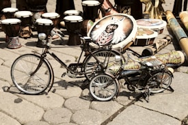 A collection of traditional drums and colorful painted musical instruments is displayed on a cobblestone surface. Two small decorative bicycles are positioned in the foreground, with intricate designs and patterns adorning their frames. The background is filled with various types of drums, including some with vibrant motifs and designs.
