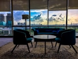 A modern interior with stylish furniture includes a central round table surrounded by two armchairs and a couch. Large windows showcase a cityscape at sunset, with vibrant clouds reflecting in the glass. A standing lamp adds to the contemporary design, and a patterned carpet covers the floor.