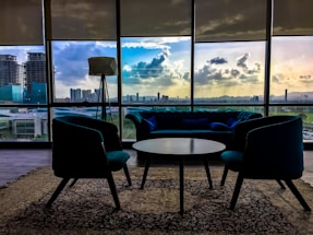 A modern interior with stylish furniture includes a central round table surrounded by two armchairs and a couch. Large windows showcase a cityscape at sunset, with vibrant clouds reflecting in the glass. A standing lamp adds to the contemporary design, and a patterned carpet covers the floor.