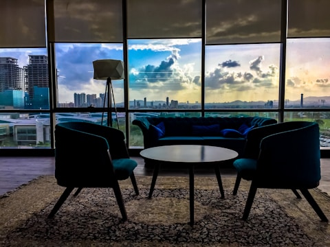 A modern interior with stylish furniture includes a central round table surrounded by two armchairs and a couch. Large windows showcase a cityscape at sunset, with vibrant clouds reflecting in the glass. A standing lamp adds to the contemporary design, and a patterned carpet covers the floor.