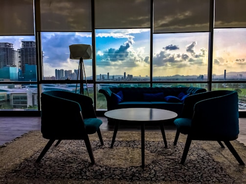A modern interior with stylish furniture includes a central round table surrounded by two armchairs and a couch. Large windows showcase a cityscape at sunset, with vibrant clouds reflecting in the glass. A standing lamp adds to the contemporary design, and a patterned carpet covers the floor.