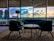 A modern interior with stylish furniture includes a central round table surrounded by two armchairs and a couch. Large windows showcase a cityscape at sunset, with vibrant clouds reflecting in the glass. A standing lamp adds to the contemporary design, and a patterned carpet covers the floor.