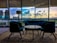 A modern interior with stylish furniture includes a central round table surrounded by two armchairs and a couch. Large windows showcase a cityscape at sunset, with vibrant clouds reflecting in the glass. A standing lamp adds to the contemporary design, and a patterned carpet covers the floor.