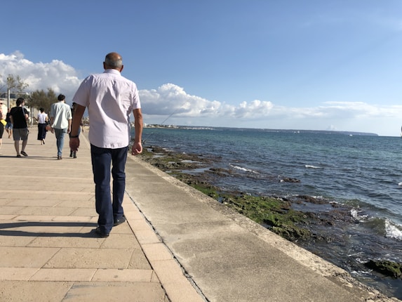 A group of people walking along a seaside promenade on a sunny day. The walkway is paved with stone tiles, and the ocean is on the right with waves gently crashing against the rocky shore. The sky is clear with a few scattered clouds.