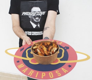 A person is holding a round tray with a colorful design featuring the word 'Mariposa'. In the center of the tray is a bowl filled with cooked chicken wings. The person is wearing a black t-shirt with a graphic of a man's face and the word 'PROGRESSIVE' written across it.
