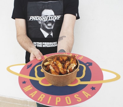 A person is holding a round tray with a colorful design featuring the word 'Mariposa'. In the center of the tray is a bowl filled with cooked chicken wings. The person is wearing a black t-shirt with a graphic of a man's face and the word 'PROGRESSIVE' written across it.