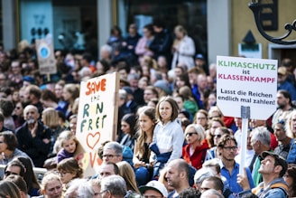 A large group of people gathered in what appears to be a protest or rally. There are signs being held up, one of which reads 'Save the RN Fret' with the word 'Love' and a heart symbol. Another sign says 'Klimaschutz ist Klassenkampf' followed by 'Lasst die Reichen endlich mal zahlen!'. The crowd seems diverse, including people of different ages and genders, and they are dressed in casual clothing.