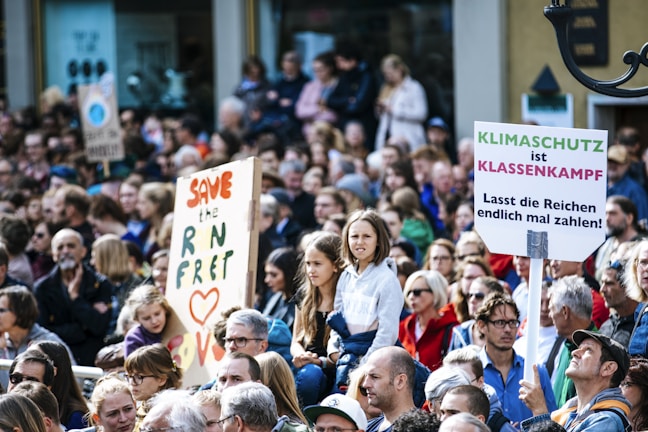 A large group of people gathered in what appears to be a protest or rally. There are signs being held up, one of which reads 'Save the RN Fret' with the word 'Love' and a heart symbol. Another sign says 'Klimaschutz ist Klassenkampf' followed by 'Lasst die Reichen endlich mal zahlen!'. The crowd seems diverse, including people of different ages and genders, and they are dressed in casual clothing.
