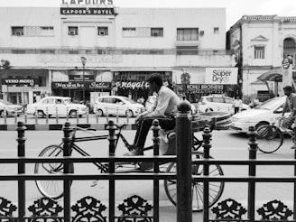 A bustling street scene features a man on a bicycle rickshaw in front of a row of shops and parked cars. The shops have signs with names like Kohli Brothers, Royal Café, and Nawabs, and a hotel named Capoor's Hotel can be seen in the background. Several people are walking along the pavement, and there are cars and motorcycles on the road. The image is in black and white, emphasizing the contrast and architectural details of the buildings.