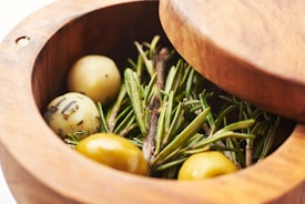 A wooden container holds several herbs and small round objects. Green sprigs of rosemary are prominently featured alongside what appear to be seasoned olives.