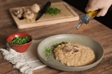 A rustic wooden table with a plate of creamy risotto garnished with mushrooms and herbs. A hand is pouring oil onto the risotto from a bottle. A red bowl filled with chopped herbs sits on a white cloth, while a cutting board with whole mushrooms, chopped herbs, and a knife is in the background.