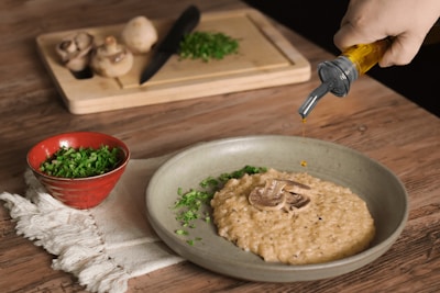 A rustic wooden table with a plate of creamy risotto garnished with mushrooms and herbs. A hand is pouring oil onto the risotto from a bottle. A red bowl filled with chopped herbs sits on a white cloth, while a cutting board with whole mushrooms, chopped herbs, and a knife is in the background.