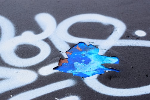 A section of black surface is covered with white spray-painted patterns, including swirling lines and abstract shapes. A portion of the surface is peeling, revealing a layer of vibrant blue paint underneath with remnants of rusted orange.