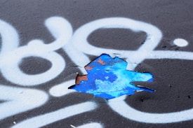 A section of black surface is covered with white spray-painted patterns, including swirling lines and abstract shapes. A portion of the surface is peeling, revealing a layer of vibrant blue paint underneath with remnants of rusted orange.
