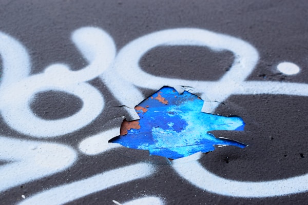 A section of black surface is covered with white spray-painted patterns, including swirling lines and abstract shapes. A portion of the surface is peeling, revealing a layer of vibrant blue paint underneath with remnants of rusted orange.