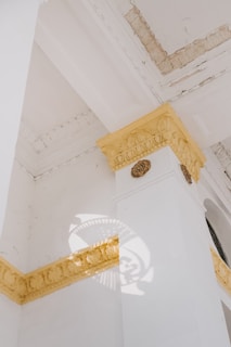 Architectural details of a building's corner with ornate moldings and intricate yellow designs. Natural light casts a decorative shadow on a white wall, highlighting the textured surface and the geometric patterns of the molding.