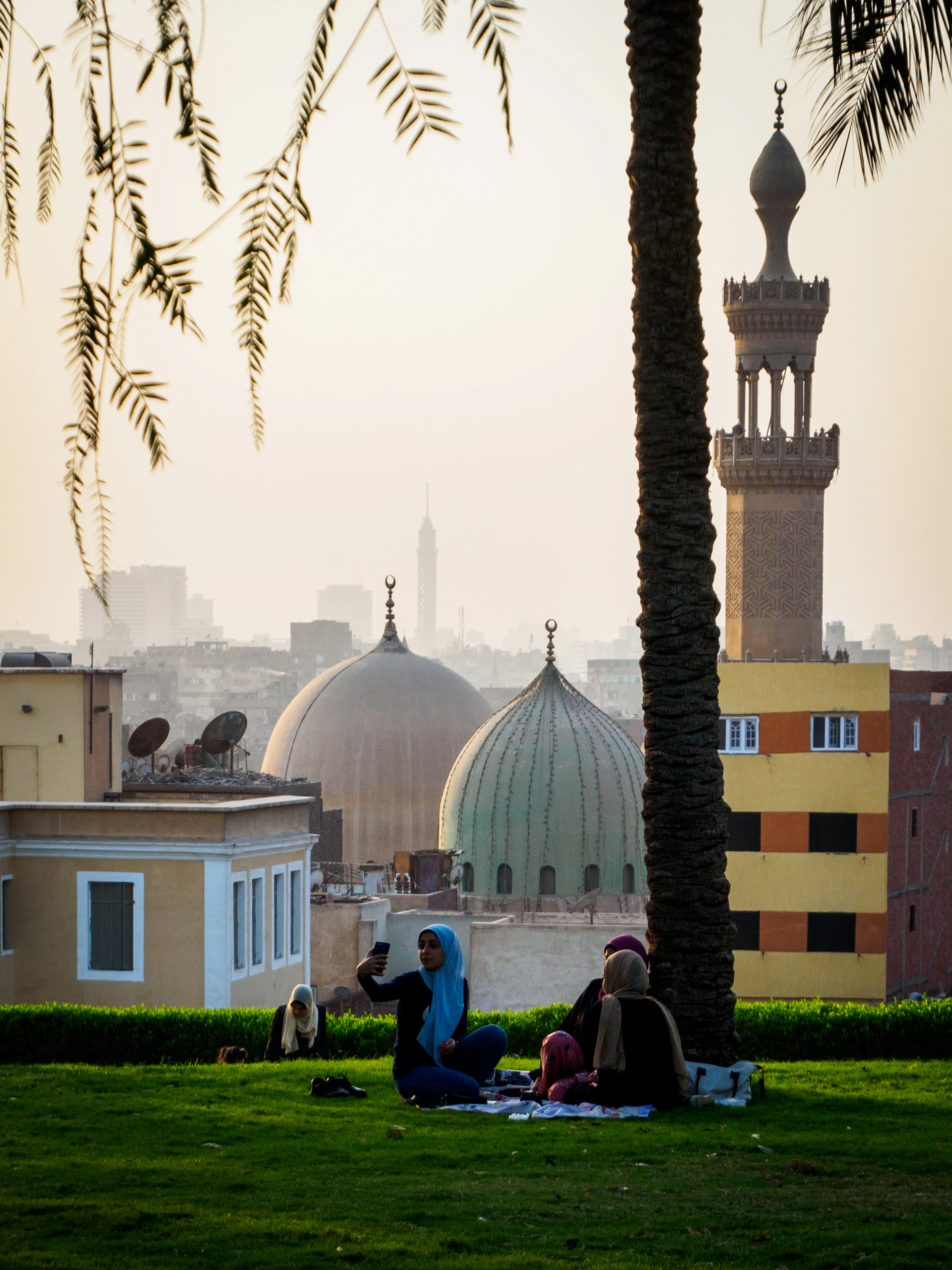Families sitting on the lawn of Al-Azhar Park in Cairo at golden hour, a palm trunk framing the domes and slender minaret of the historic city as the hazy skyline fades behind them.