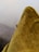 A close-up view of a small insect, possibly a mosquito, resting on the corner of a mustard-colored fabric cushion. The texture of the cushion is visible, with a grainy and slightly fuzzy surface. In the background, there is a soft gray fabric, possibly another cushion or a piece of furniture, against a plain light-colored wall.