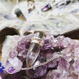 A collection of amethyst crystals and a glass vial pendant with silver accents are displayed on top of the crystals. There is a delicate chain with purple and blue beads, contributing to the intricate arrangement. In the background, there is a slightly blurred reflective surface with additional crystal pendants.