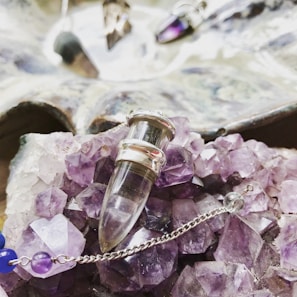 A collection of amethyst crystals and a glass vial pendant with silver accents are displayed on top of the crystals. There is a delicate chain with purple and blue beads, contributing to the intricate arrangement. In the background, there is a slightly blurred reflective surface with additional crystal pendants.