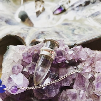 A collection of amethyst crystals and a glass vial pendant with silver accents are displayed on top of the crystals. There is a delicate chain with purple and blue beads, contributing to the intricate arrangement. In the background, there is a slightly blurred reflective surface with additional crystal pendants.