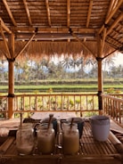 A tropical outdoor setting features a bamboo structure with a thatched roof overlooking lush green fields and palm trees. In the foreground, a wooden table holds several glass jars filled with frothy drinks, each with a straw, alongside a ceramic mug.