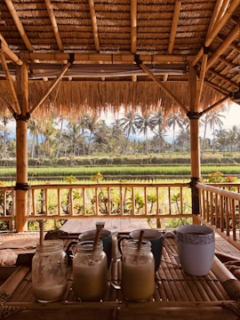 A tropical outdoor setting features a bamboo structure with a thatched roof overlooking lush green fields and palm trees. In the foreground, a wooden table holds several glass jars filled with frothy drinks, each with a straw, alongside a ceramic mug.