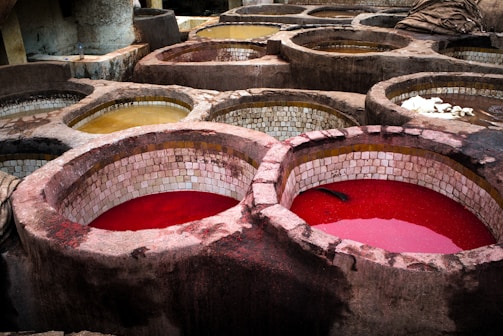 Circular stone vats filled with various colored liquids, predominantly red and yellow, set in a traditional tannery environment. The vats have tiled interiors and are used for dyeing or tanning processes.