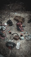 A collection of items scattered on dry grass includes a hookah, a glass bottle, a couple of soda bottles, a crumpled brown jacket, a red and black checkered shirt, a brown bag, crumpled foil wrappers, and a cup. The scene has a rustic, disorganized feel.