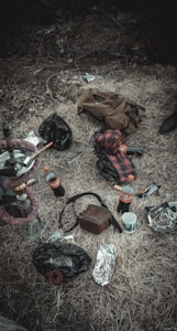 A collection of items scattered on dry grass includes a hookah, a glass bottle, a couple of soda bottles, a crumpled brown jacket, a red and black checkered shirt, a brown bag, crumpled foil wrappers, and a cup. The scene has a rustic, disorganized feel.