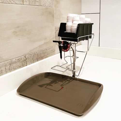 A plastic container holding stacks of small paper cups is mounted on a metal stand by a tiled wall. Below it, a large plastic tray is placed on a white surface, likely to catch any drips from the tap on the container.