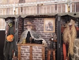 Scene depicting a Halloween-themed setup with three spooky figures resembling witches or scarecrows in front of a faux brick wall. The central figure, dressed in tattered, web-covered clothing, stands behind a rustic wooden bar with a large bottle and goblet atop. To the left, a green-faced figure holds a carved pumpkin. To the right, a rustic scarecrow-like figure has straw for hair. Above them, a picture of a haunted house adds to the eerie atmosphere.