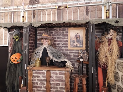 Scene depicting a Halloween-themed setup with three spooky figures resembling witches or scarecrows in front of a faux brick wall. The central figure, dressed in tattered, web-covered clothing, stands behind a rustic wooden bar with a large bottle and goblet atop. To the left, a green-faced figure holds a carved pumpkin. To the right, a rustic scarecrow-like figure has straw for hair. Above them, a picture of a haunted house adds to the eerie atmosphere.