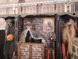 Scene depicting a Halloween-themed setup with three spooky figures resembling witches or scarecrows in front of a faux brick wall. The central figure, dressed in tattered, web-covered clothing, stands behind a rustic wooden bar with a large bottle and goblet atop. To the left, a green-faced figure holds a carved pumpkin. To the right, a rustic scarecrow-like figure has straw for hair. Above them, a picture of a haunted house adds to the eerie atmosphere.