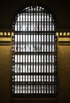 A large, arched window with a grid pattern of metal bars and frosted glass panels. The structural elements feature ornate decorative designs typical of classic architecture. Silhouettes of people are visible through the glass, suggesting movement beyond the window.